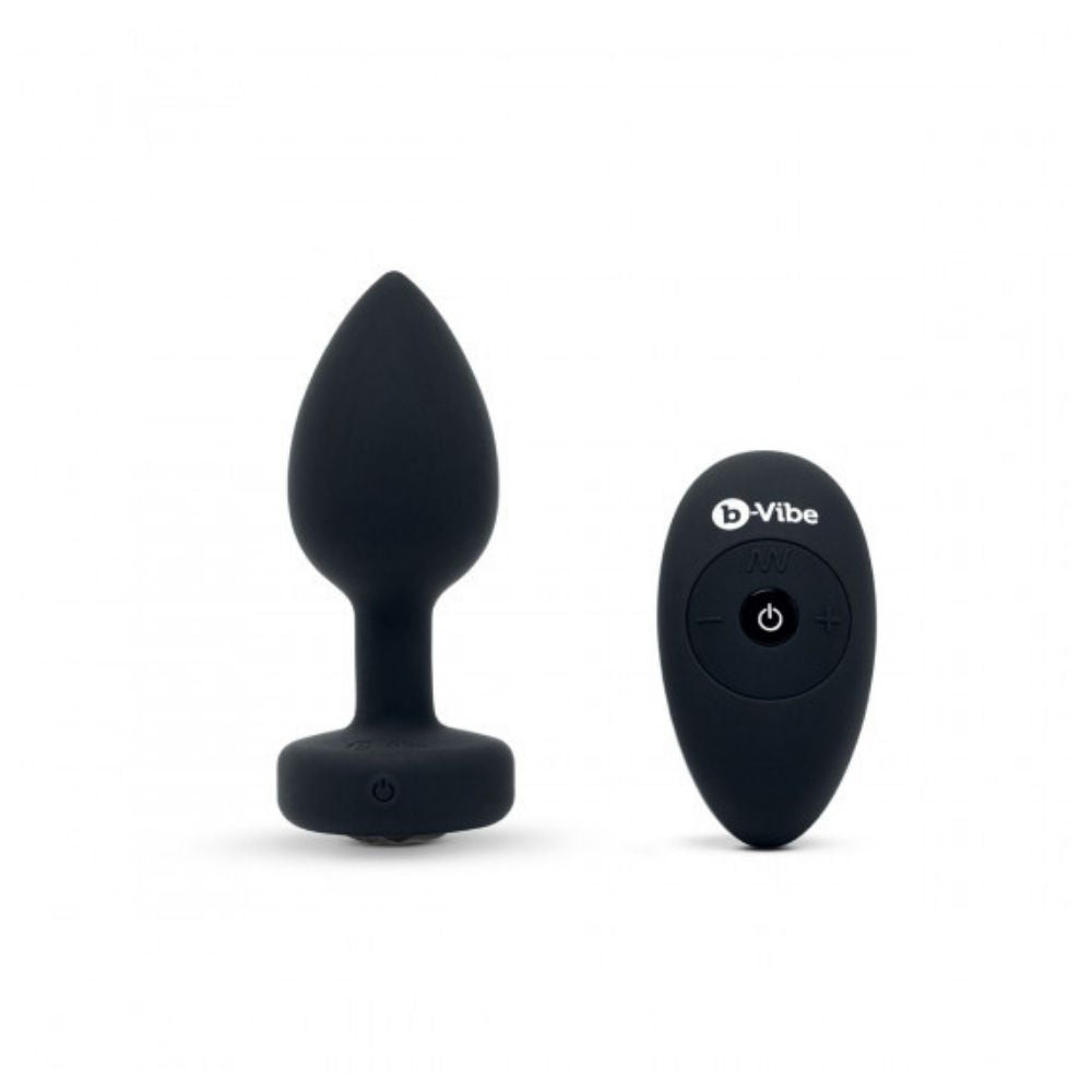 Black B-Vibe Vibrating Jewel Plug Medium/Large standing upright on its base beside the remote control it comes with